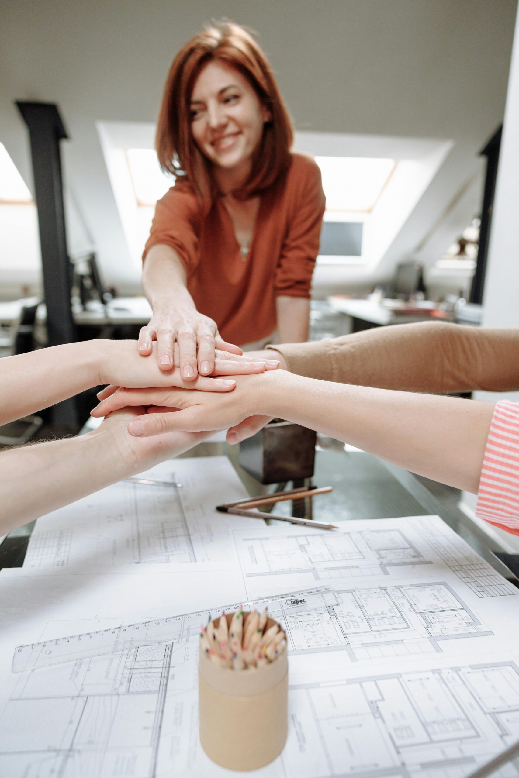A group of professionals joining hands over desk plans in a modern office, symbolizing teamwork.